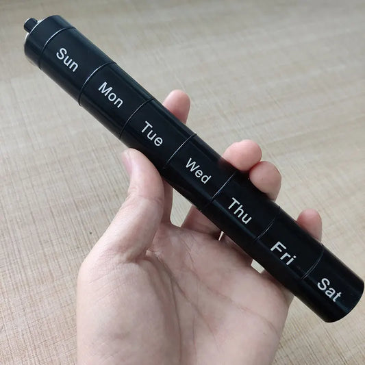Black travel pill organizer with days of the week labels held in a hand on a wooden surface